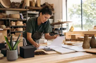 Middle-aged woman entrepreneur reviewing paperwork in a sunlit artisan workshop, surrounded by tools and business materials, absorbed in her work.