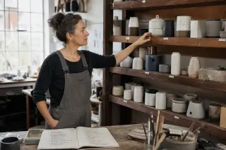 Entrepreneuse en blouse de travail observant des prototypes sur une étagère, carnet ouvert rempli de notes sur un établi, lumière naturelle douce dans un atelier artisanal.